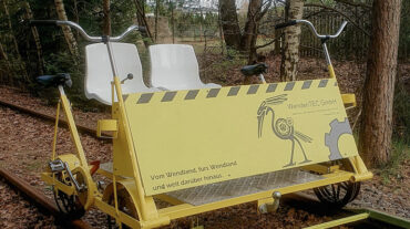 Dieses Bild zeigt eine gelbe Draisine auf Schienen. Die Draisine kann mit vier Personen gefahren werden. Die beiden äußeren Sitze können mit Pedalen wie an einem Fahrrad. Dazwischen sind zwei Schalensitze angebraucht. Auf der Vorderseite der Draisine ist zu lesen: Vom Wendland, fürs Wendland und weit darüber hinaus, Wenden Tec GmbH. Zwischen den Gleisen liegt Laub.