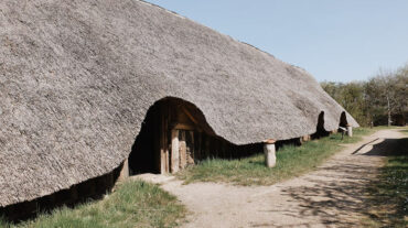 Dieses Bild zeigt ein Langhaus in dem Archeologischen Zentrum in Hitzacker. Das Dach ist mit Reet gedeckt und reicht bis fast auf den Boden. Entlang des Hauses führt ein Sandweg, von dem es drei Eingänge zu Haus gibt.