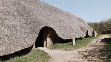 langhaus Dieses Bild zeigt ein Langhaus in dem Archeologischen Zentrum in Hitzacker. Das Dach ist mit Reet gedeckt und reicht bis fast auf den Boden. Entlang des Hauses führt ein Sandweg, von dem es drei Eingänge zu Haus gibt.