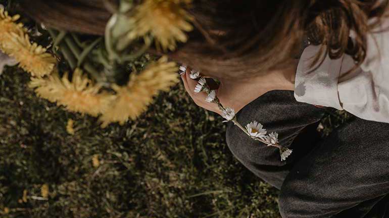 Dieses Bild zeigt ein Kind, welches einen Blumenkranz aus Gänseblümchen flechtet. Es hat einen Kranz aus Löwezahnblumen auf dem Kopf. Das Bild ist von oben über die Schulter des Kindes aufgenommen.