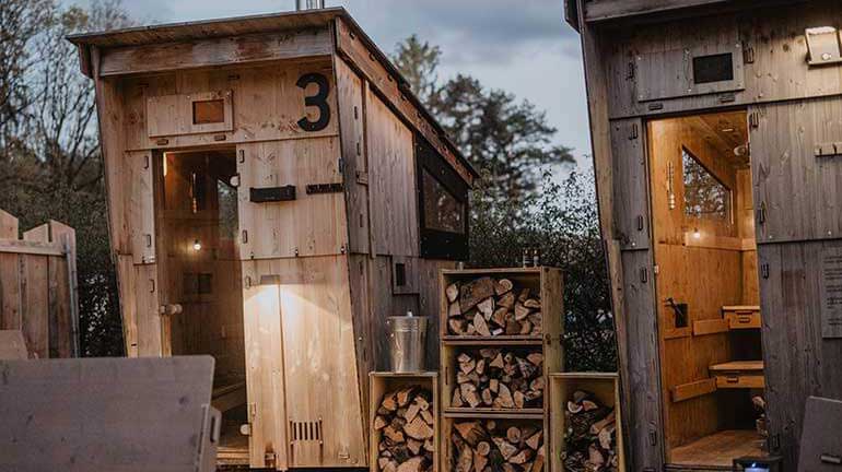 Dieses Bild zeigt zwei Saunen aus Holz mit beleuchteten Eingängen. Zwischen den Saunen ist Brennholz in Holzkisten gestapelt und oben drauf steht ein Aufgusseimer. In den Saunen sieht es hell und sehr gemütlich aus.