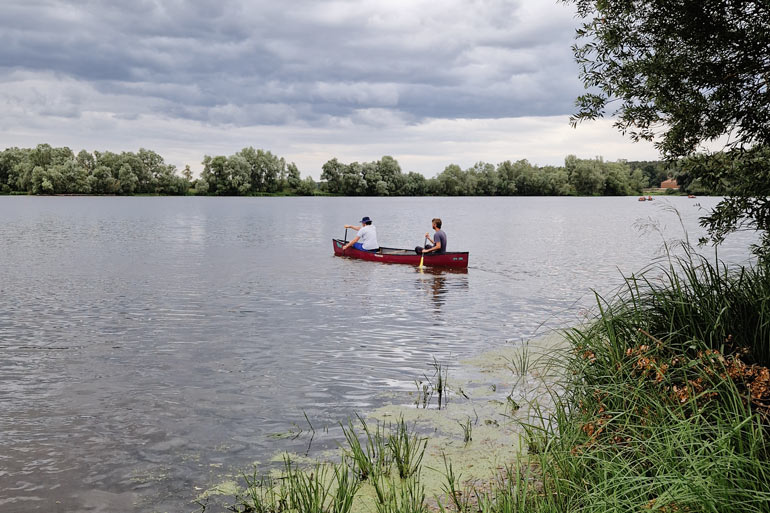 Dieses Bild zeigt einen See, auf dem zwei Personen in einem Kanu sitzen. Sie machen eine Kanutour auf dem Hitzacker See. Im Vordergrund ist ds Ufer zu sehen, welches von Schilf bewachsen ist. Das gegenüberliegende Ufer ist voll mit Bäumen. Der Himmel ist bewölkt.