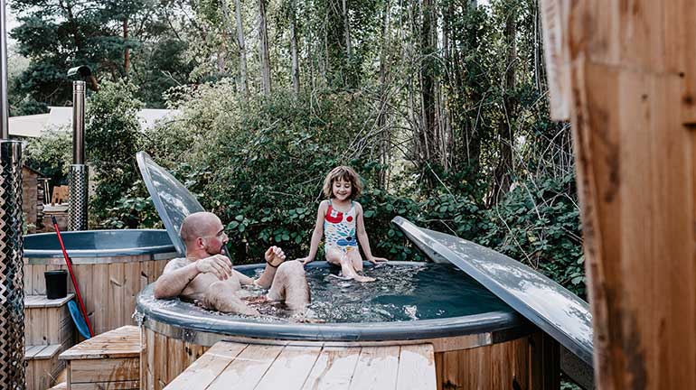 Dieses Bild zeigt einen Badezuber aus Holz mit einem Erwachsenen und einem Kind, die darin baden. Das Kind sitzt auf dem Rand. Die Abdeckung lehnt am Badezuber.