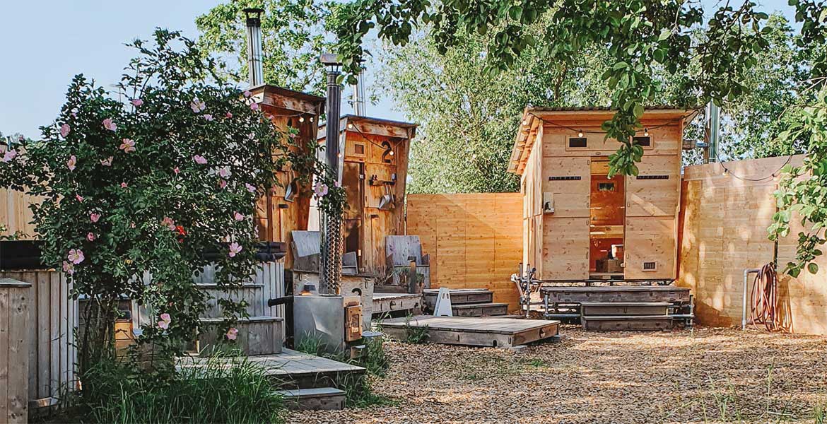 Dieses Bild zeigt eine große Holzsauna unter Bäumen. Links daneben stehen zwei weitere kleine Saunen mit Holzterrassen davor. Zwischen den Saunen ist ein Sichtschutz aus Holz. Links neben den Saunen stehen zwei Badezuber, die teilweise von einer wilden Rose verdeckt werden.