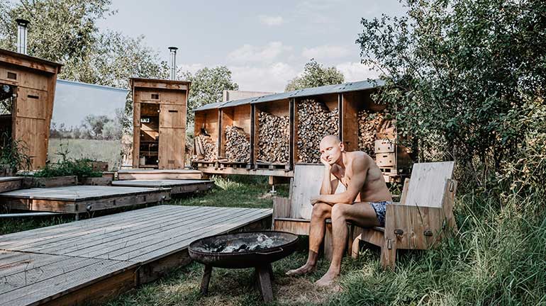 Dieses Bild zeigt einen Erwachsenen, der auf einem Sessel aus Holz im Freien sitzt. Vor Ihm ist eine Holzterrrasse und eine Feuerschale. Im Hintergrund ist Brennholz aufgestappelt neben Eingängen zu zwei Saunen aus Holz.